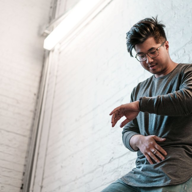 Image description: A person leans against a wall, checking their watch.
