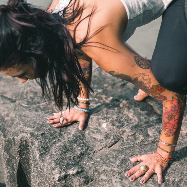 Close up photo of a person practicing crow pose, Bakasana, on a rock near water.