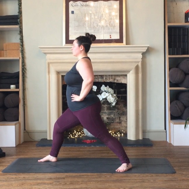 Sarah holds a Warrior 1 posture with her hands on her hips